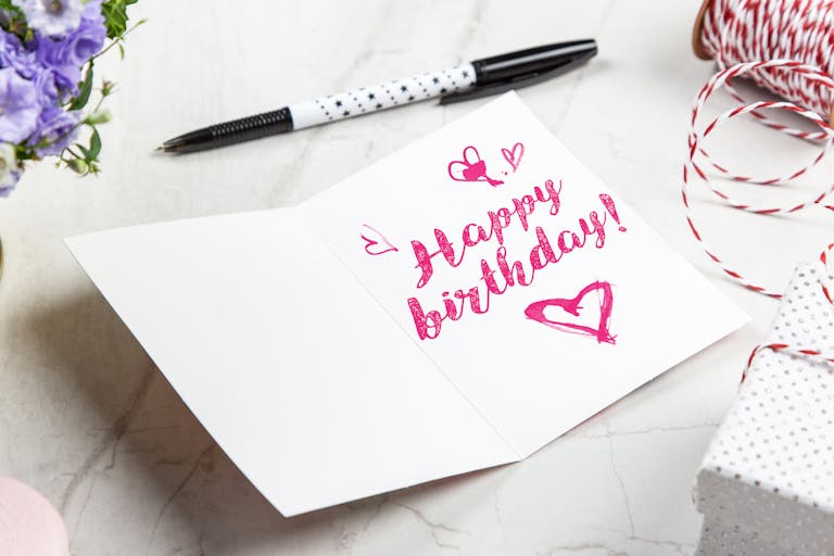 A vibrant birthday card with heart illustrations, surrounded by a pen, flowers, and gift wrap.
